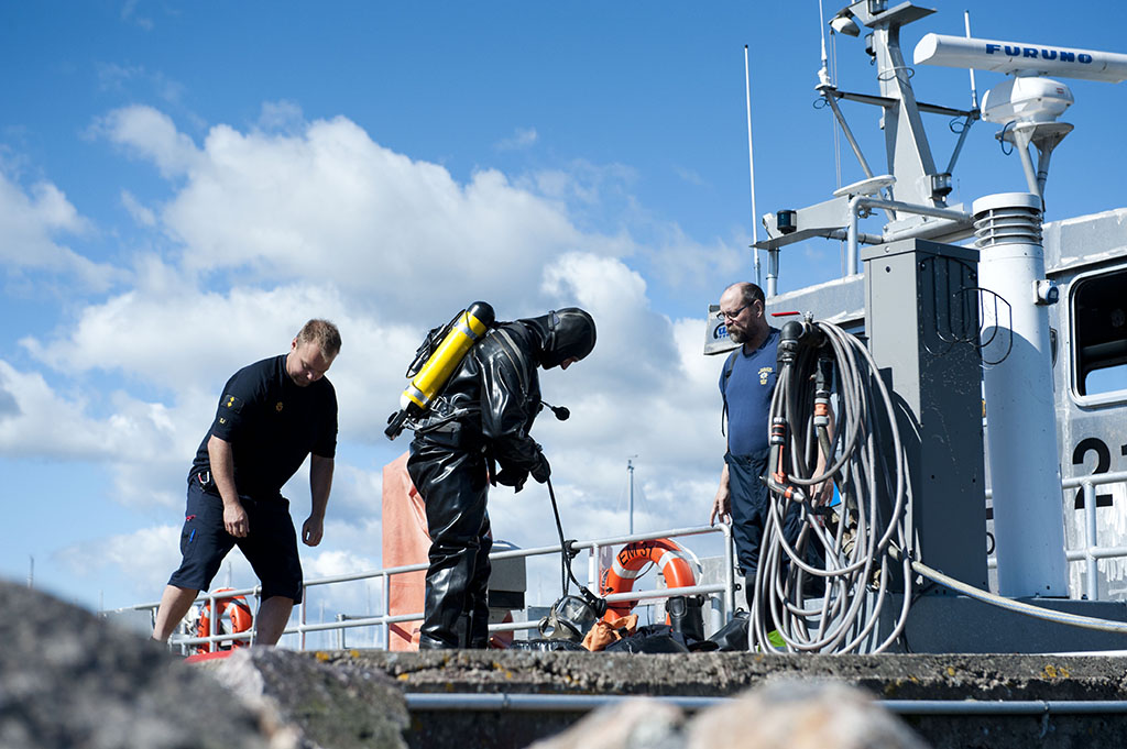 Paloesimies Sami Jauhiainen auttaa sukeltaja Juho Marjasta kuivapuvun kanssa Nuottaniemen venesatamassa. Oikealla ylipalomies Kauko Vasse.